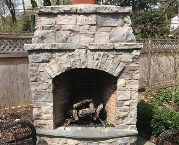 Rustic stone outdoor fireplace with an arched firebox and chimney, built by Top Hat Masonry Repair, set in a cozy backyard patio with wooden fencing